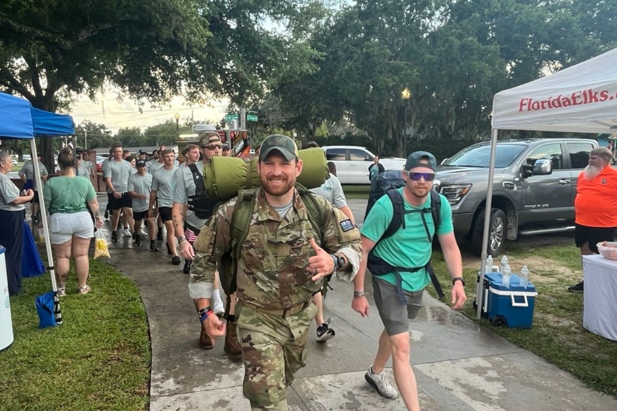 Grass to the left, two men walking on a gray concrete pathway, the one on the left dressed in combat fatigues, the man on the right in a green shirt, both wearing backpacks, with vehicles and tents to their right, and with people in the background, walking under a canopy of trees.