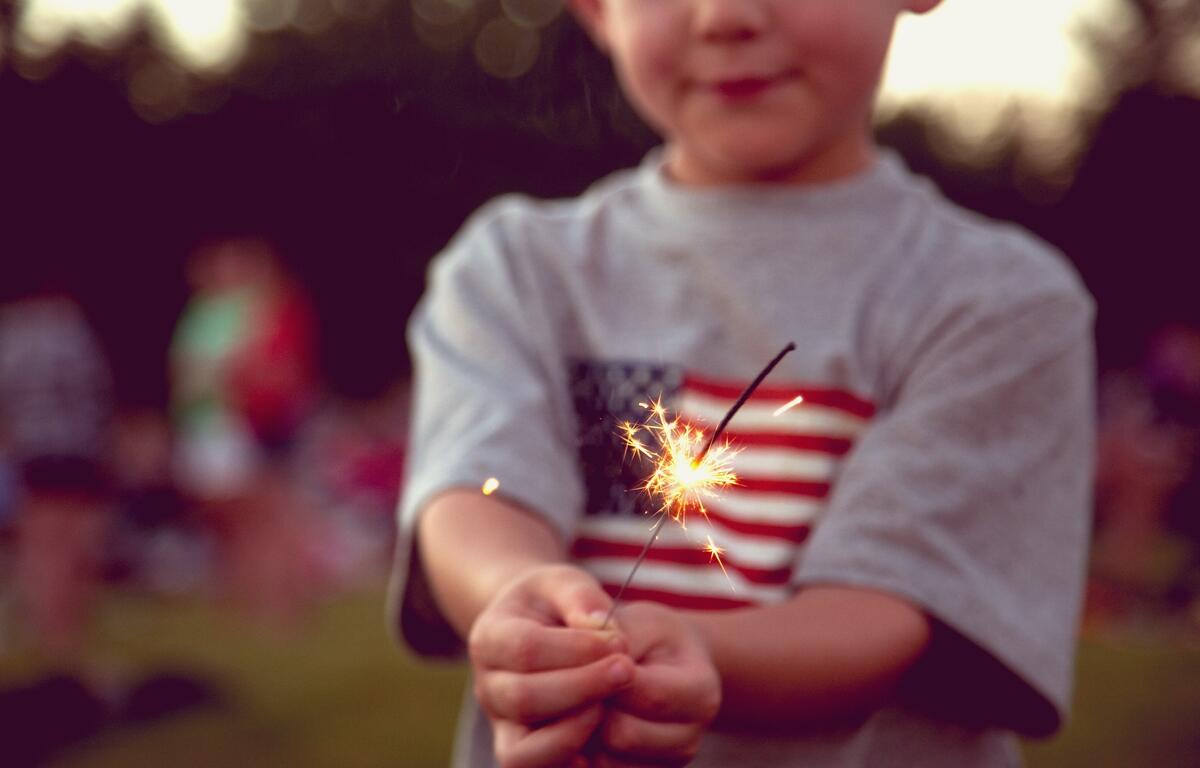 A child holding a lit sparkler, wearing a shirt with an American flag design, with a blurred outdoor background.