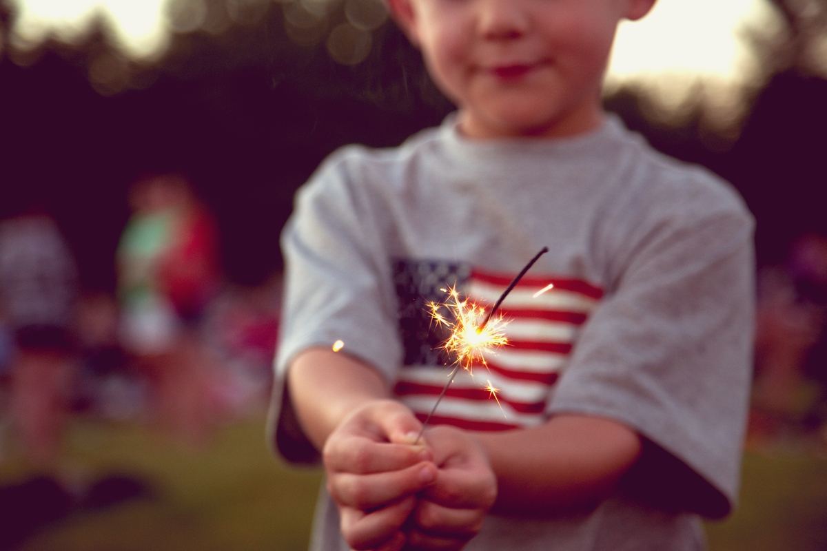 A child holding a lit sparkler, wearing a shirt with an American flag design, with a blurred outdoor background.