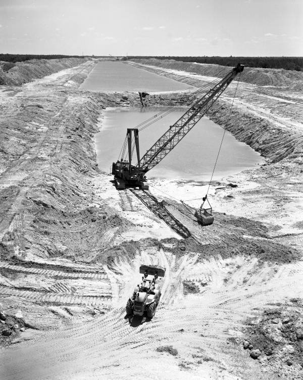 A photograph showing a view of the partially excavated canal near Ocala, Florida. Large mounds of earth and a wide trench cut through a forested area, with construction equipment actively digging.