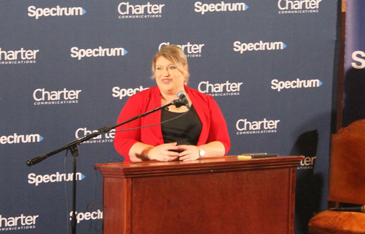 A well-dressed woman standing behind a podium with a microphone to her left in front of a blue background with lettering.