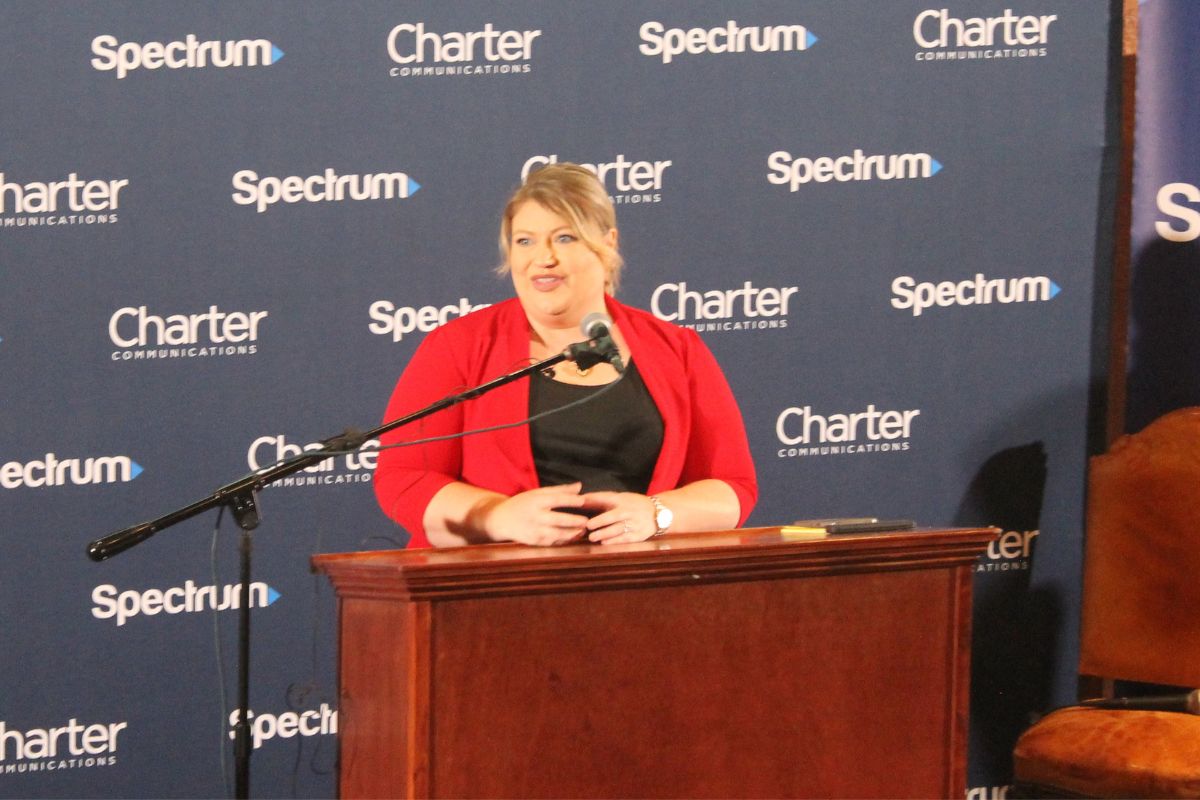 A well-dressed woman standing behind a podium with a microphone to her left in front of a blue background with lettering.