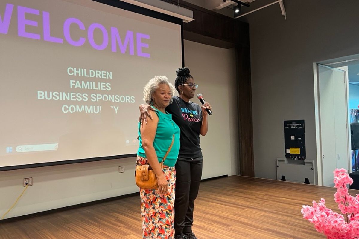 Two women on a wooden floor stage, standing in front of a screen with writing on it, the woman to the right has her arm around the woman to her left's shoulder, with a floral bouquet in the forefront.