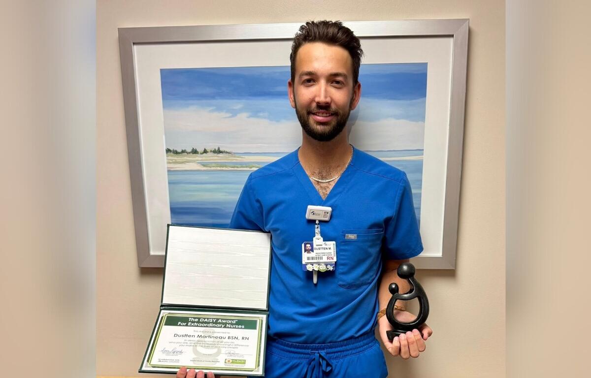 Dustten Martineau in blue scrubs holding his DAISY Award certificate and statue in front of a framed beach painting.