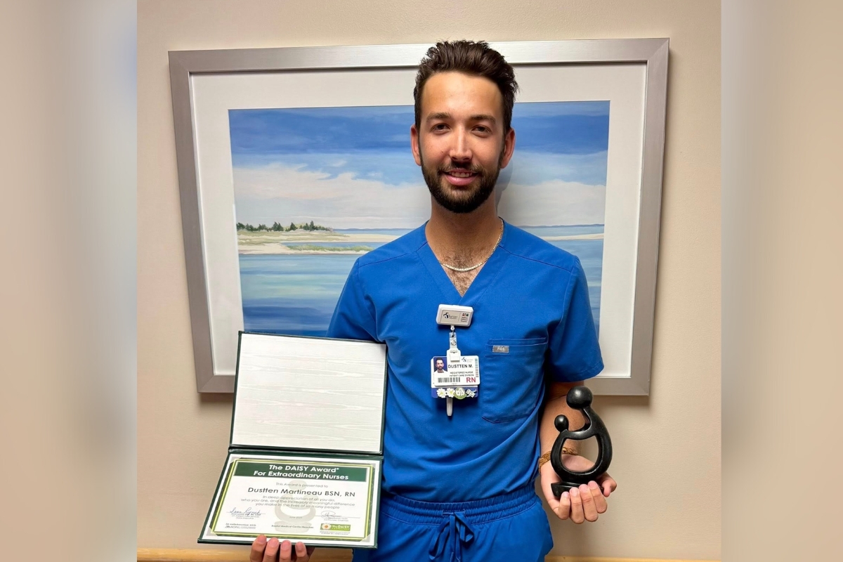 Dustten Martineau in blue scrubs holding his DAISY Award certificate and statue in front of a framed beach painting.