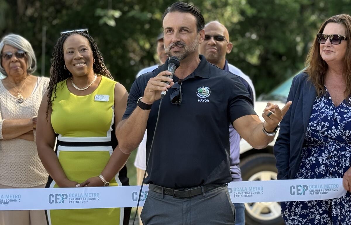 Ocala Mayor Ben Marciano speaks into a microphone at a ribbon cutting event, standing in front of a group of attendees.