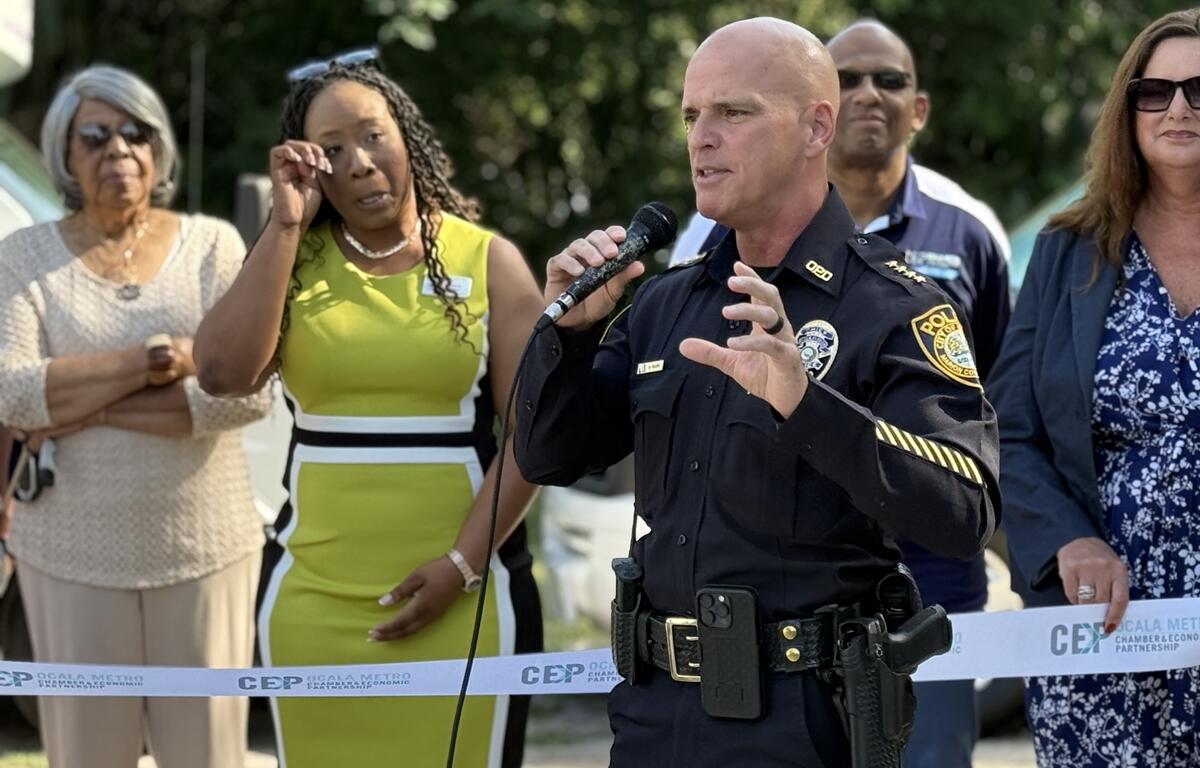 Ocala Police Chief Mike Balken speaks into a microphone at a ribbon cutting event with attendees standing nearby.