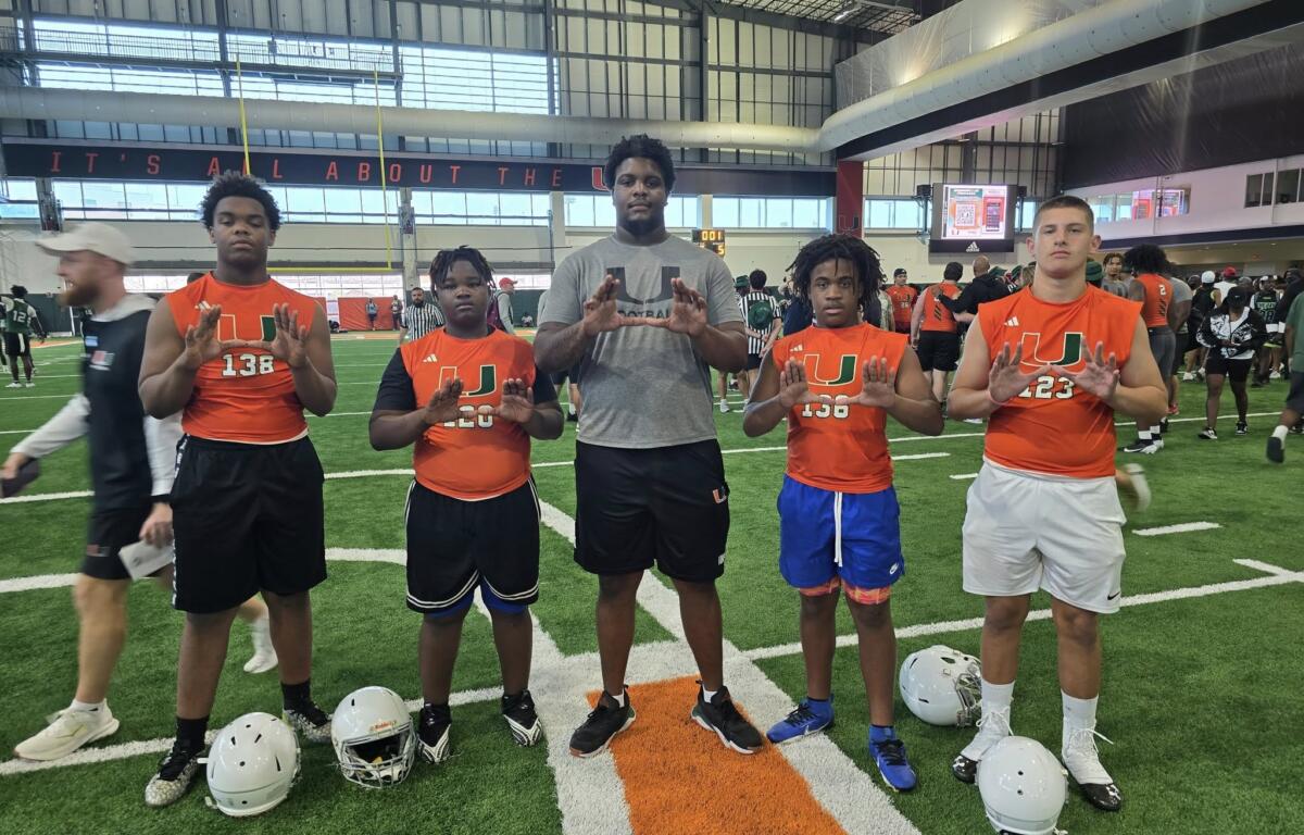 A group of young athletes in orange Miami jerseys and a coach pose together on the indoor field at the Empowered Linemen Academy, proudly holding up the signature “U” hand gesture.