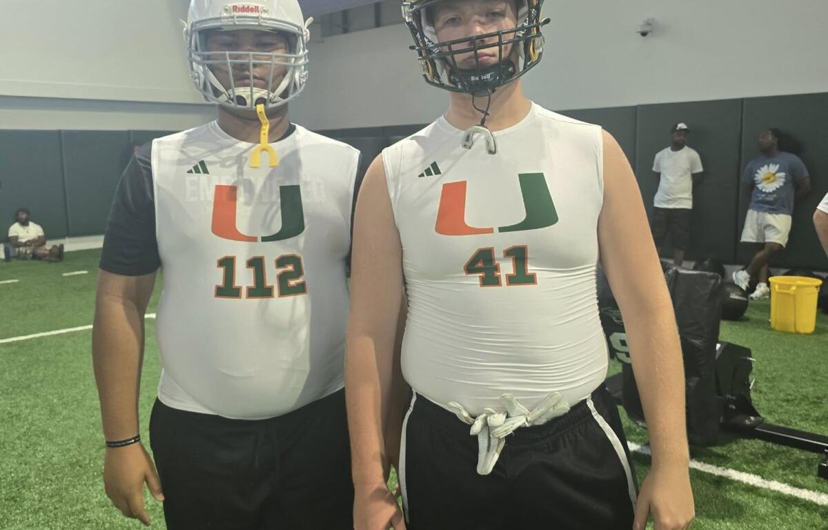 Two young linemen in white Empowered Linemen Academy jerseys and helmets pose confidently inside an indoor training facility, standing beneath a large Adidas logo after a day of drills and development.
