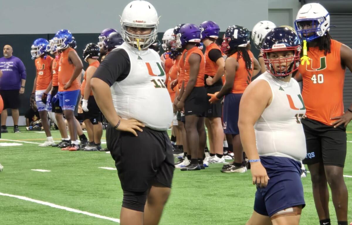 Two young linemen in white Miami football jerseys stand on the field during a drill session at the Empowered Linemen Academy, with other athletes and coaches preparing in the background.