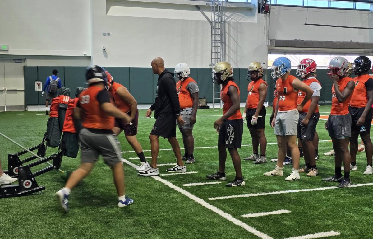 A group of young football players wearing orange sleeveless jerseys and helmets wait in line during a lineman training drill, where one participant is shown striking a practice sled under the guidance of a coach.