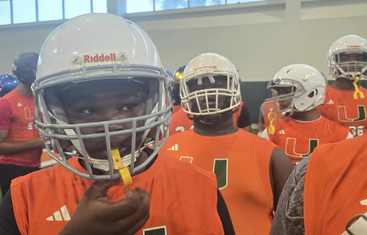 Young football players in orange Miami jerseys and white helmets stand focused and ready during a drill at the Empowered Linemen Academy, with one adjusting his mouthguard.