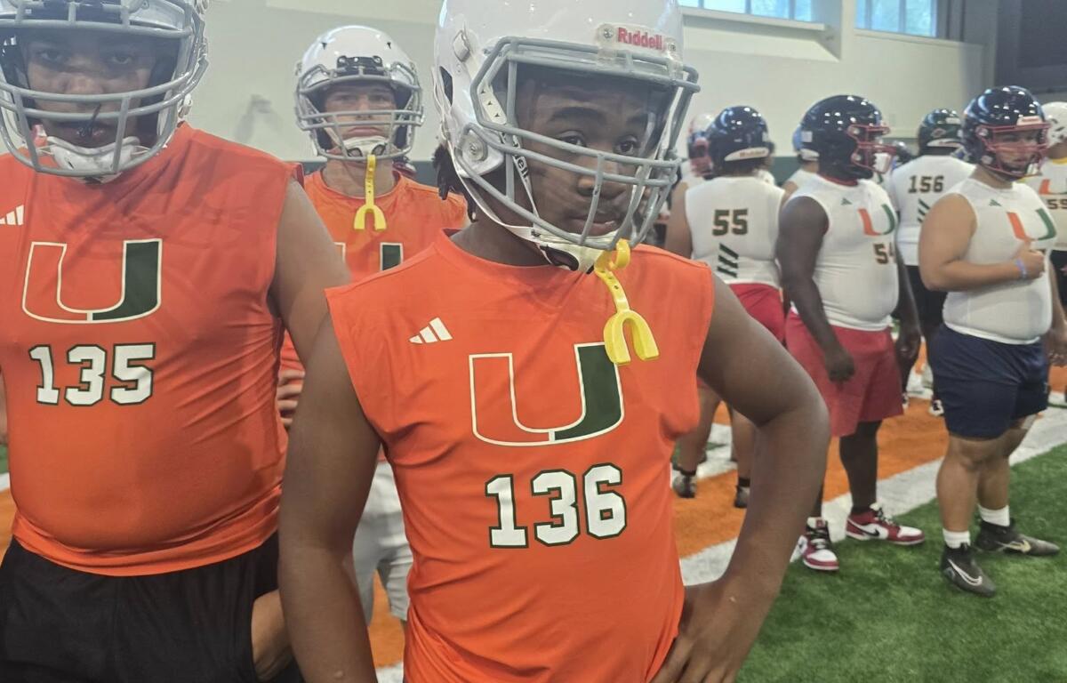 A young football player wearing a white helmet, orange "U" sleeveless jersey numbered 136, and blue athletic shorts stands confidently on an indoor turf field. Behind him are other similarly dressed athletes preparing for drills, suggesting a skills camp or training event at the University of Miami.
