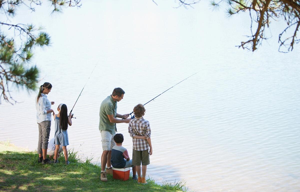 Family with young children fishing from the grassy shore of a freshwater lake under tree shade.