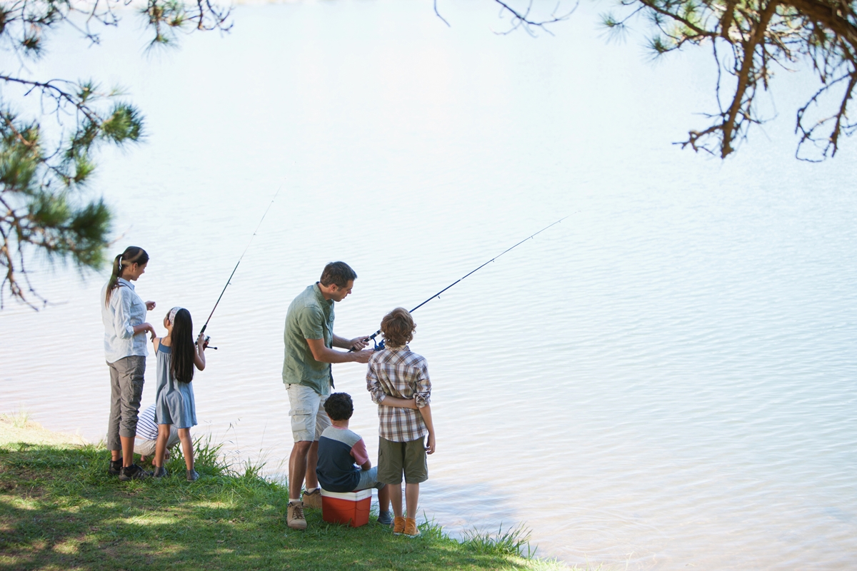 Family with young children fishing from the grassy shore of a freshwater lake under tree shade.