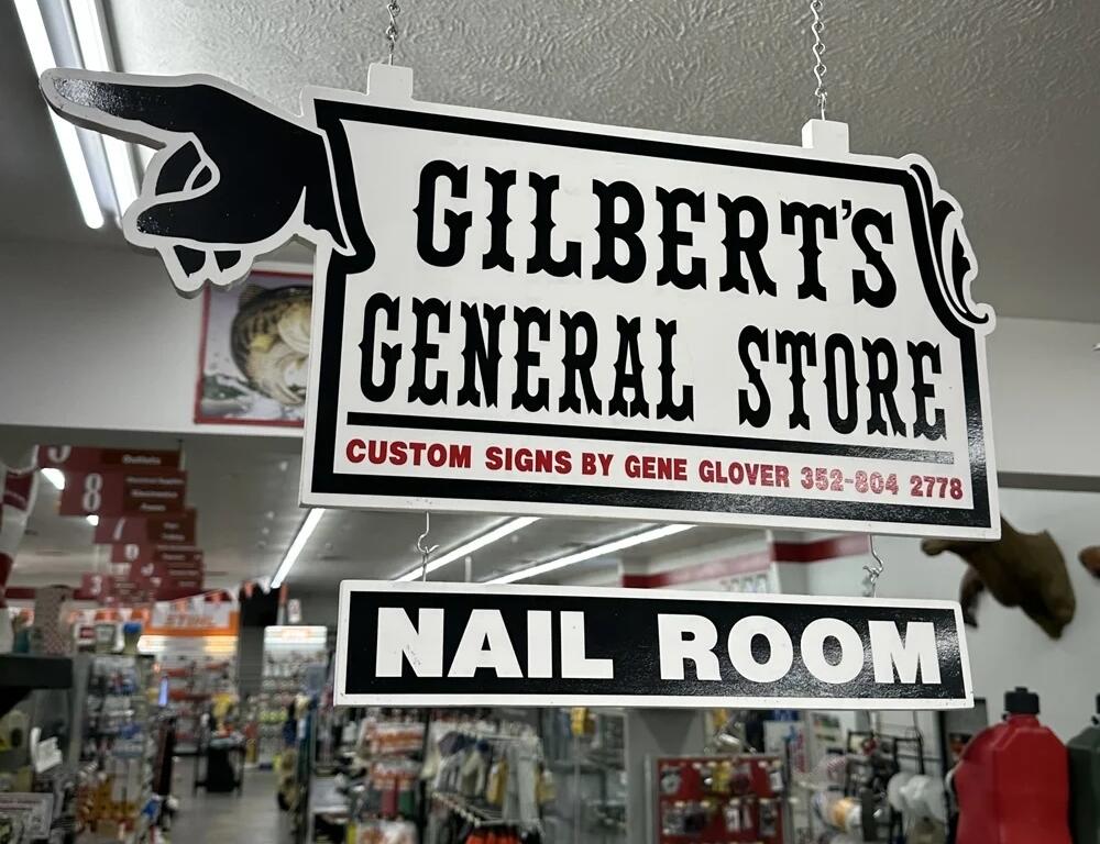Hanging sign inside Gilbert’s General Store reads “Nail Room” with a pointing hand and credits local sign maker Gene Glover. Aisles of hardware supplies are visible in the background.
