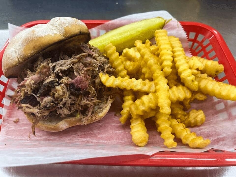 Close-up of a deli meal featuring a pulled pork sandwich on a bun, crinkle-cut French fries, and a pickle spear in a red plastic basket with parchment paper.