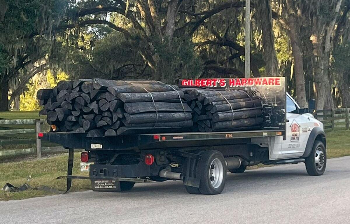 Flatbed Gilbert’s Hardware truck loaded with black wooden fence posts driving down a tree-lined rural road in Marion County.