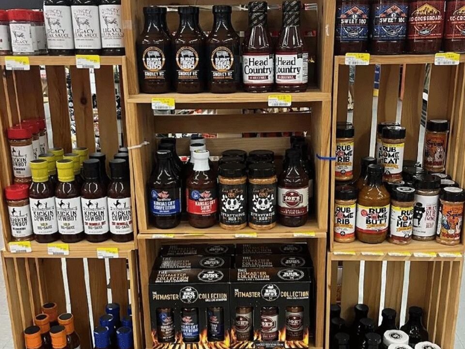 Wooden shelves filled with various barbecue sauces, spice rubs, and grilling seasonings at Gilbert’s Hardware, including brands like Kickin’ Cowboy and Head Country.