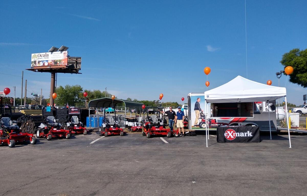 Outdoor display of Exmark zero-turn mowers with staff under a white vendor tent, surrounded by balloons and promotional signage.