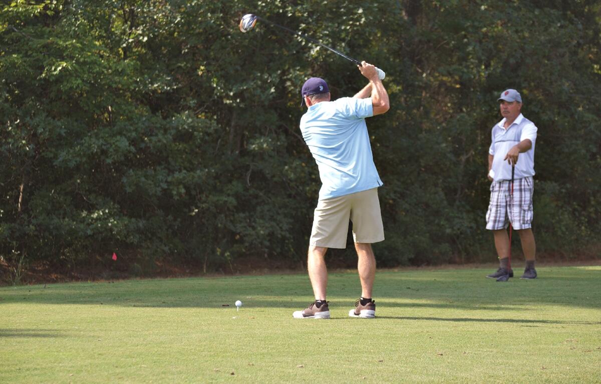 Two men golfing, one mid-swing at the tee and the other watching nearby on a sunny day.