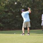Two men golfing, one mid-swing at the tee and the other watching nearby on a sunny day.