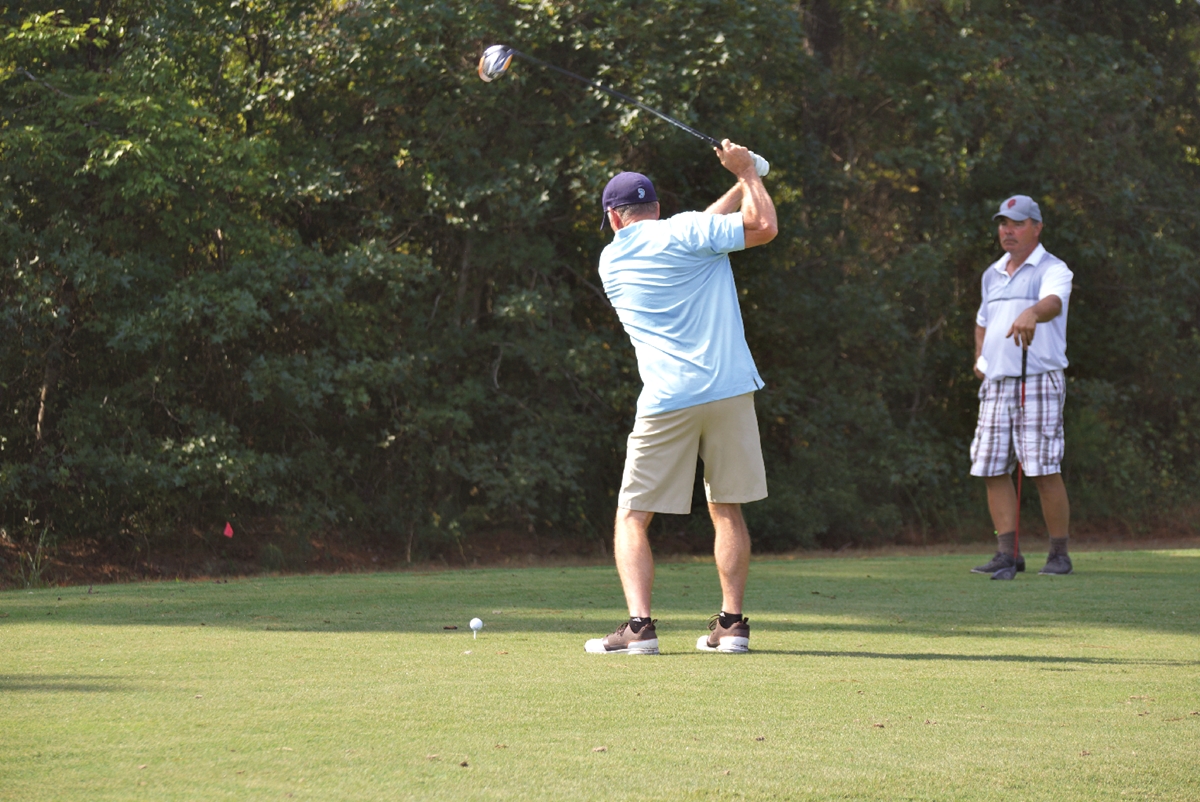 Two men golfing, one mid-swing at the tee and the other watching nearby on a sunny day.