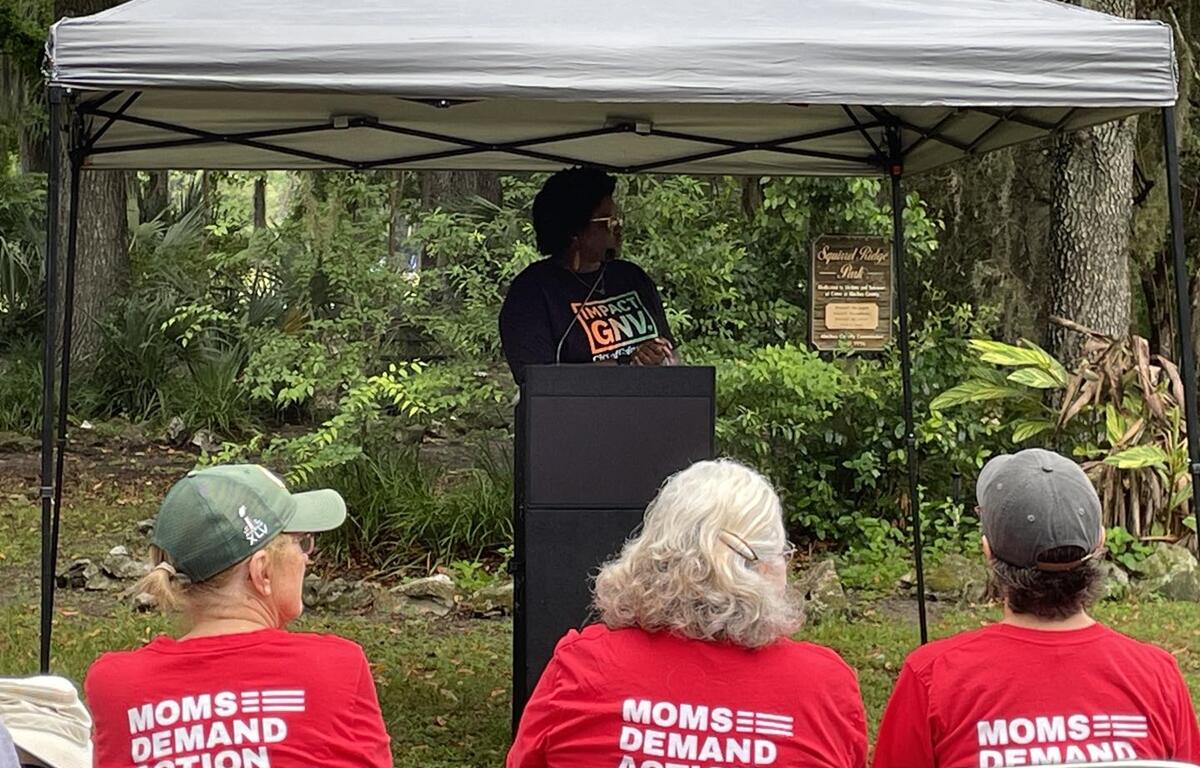 Woman speaks at podium under canopy during gun violence memorial event, with Moms Demand Action members in red shirts seated in front at Squirrel Ridge Park in Gainesville.