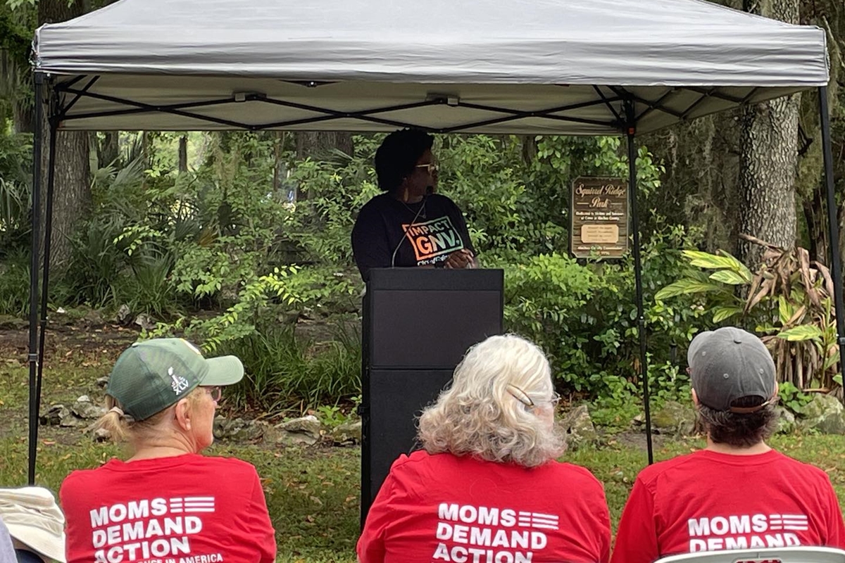 Woman speaks at podium under canopy during gun violence memorial event, with Moms Demand Action members in red shirts seated in front at Squirrel Ridge Park in Gainesville.