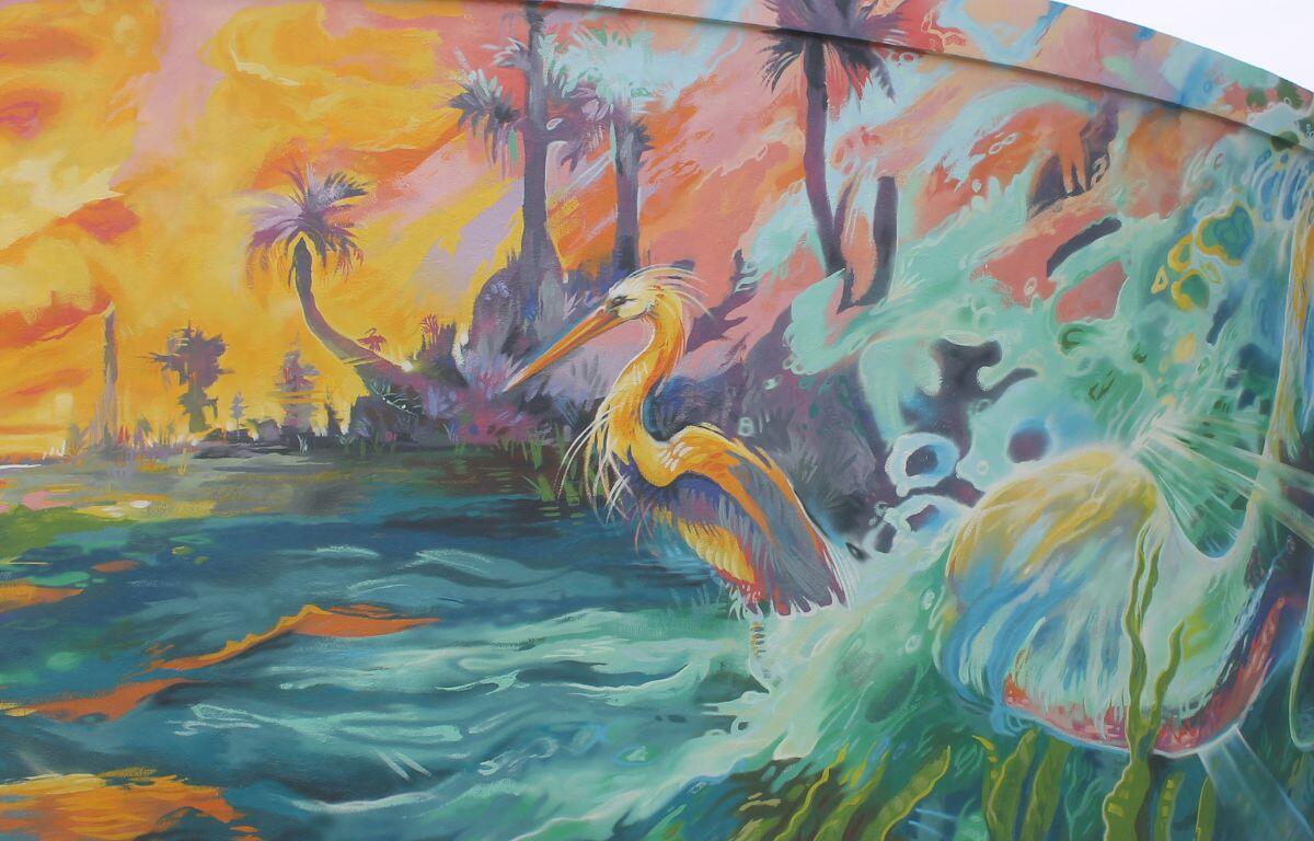 A large bird sits on the edge of the water to the left, with a sunset and palms in the background, with grass to the right.