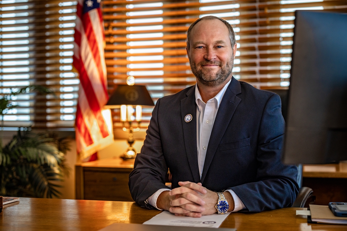 Gregory C. Harrell seated at his desk in an office with an American flag and blinds in the background.