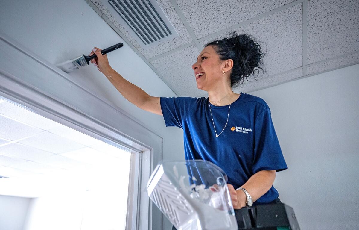 Woman painting a white door frame while standing on a ladder inside Heart of the Park.