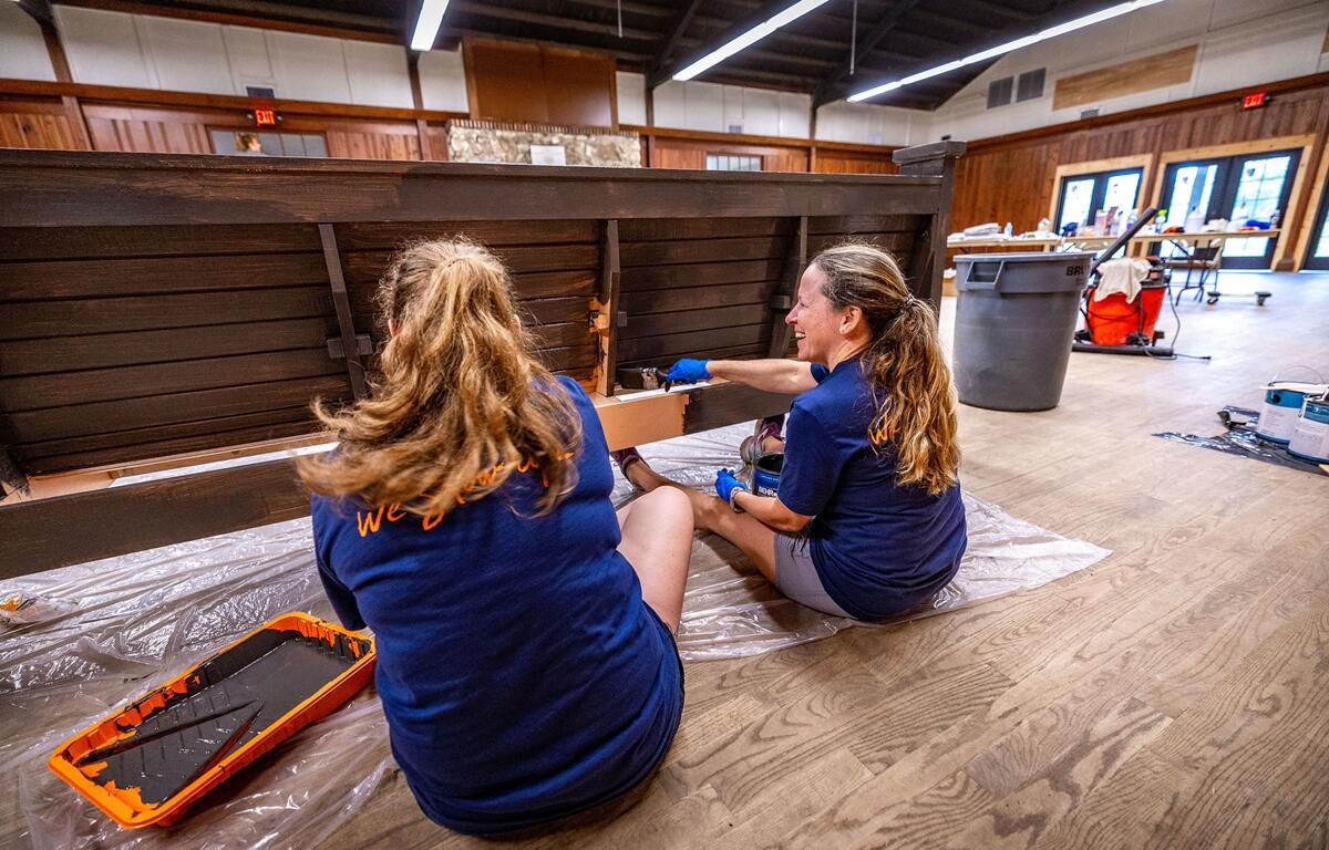 Two women in “We show up” shirts painting a bench inside a wooden community hall.