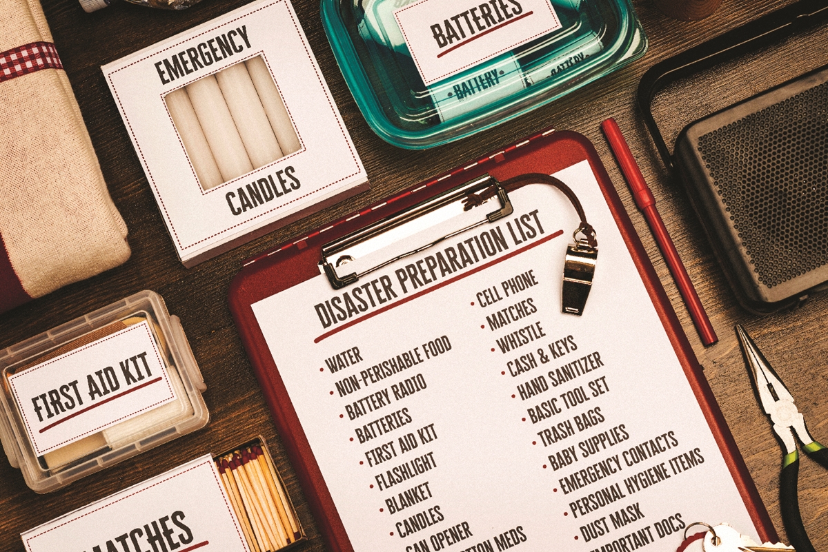 Hurricane preparedness kit with labeled items including candles, batteries, first aid kit, matches, and a disaster supply checklist on a clipboard.