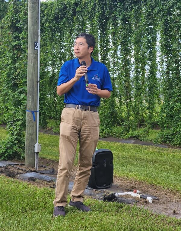 Shinsuke Agehara holds a microphone while speaking outdoors in front of tall hop vines.