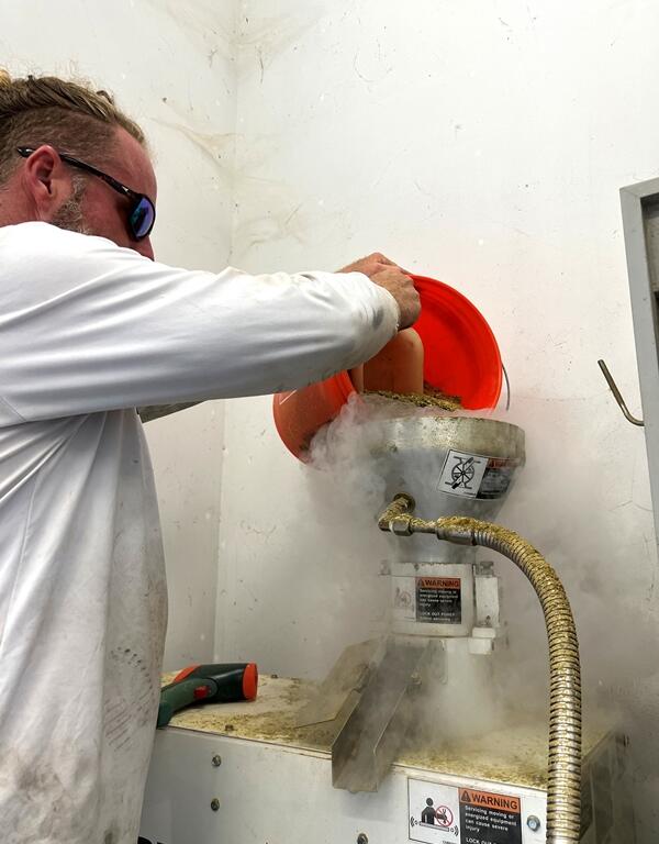 Man pours hops from an orange bucket into a steaming pelletizing machine inside a processing room.