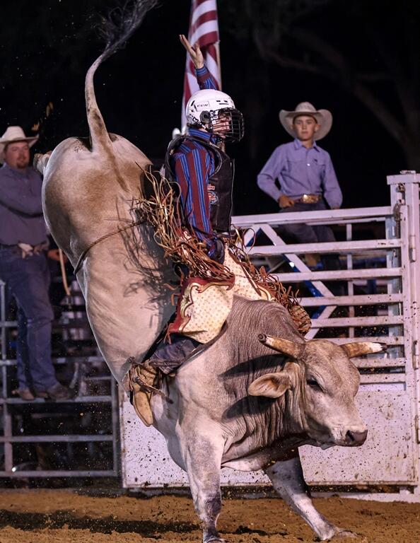 Bull rider in protective gear rides a bucking bull during a nighttime rodeo event with spectators watching from behind the gate.