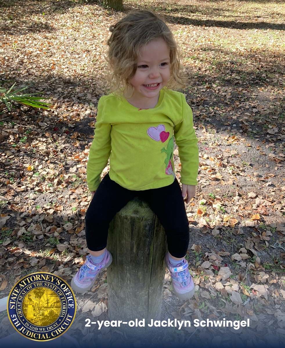 Smiling toddler Jacklyn Schwingel, age 2, sits on a wooden post outdoors surrounded by fallen leaves.
