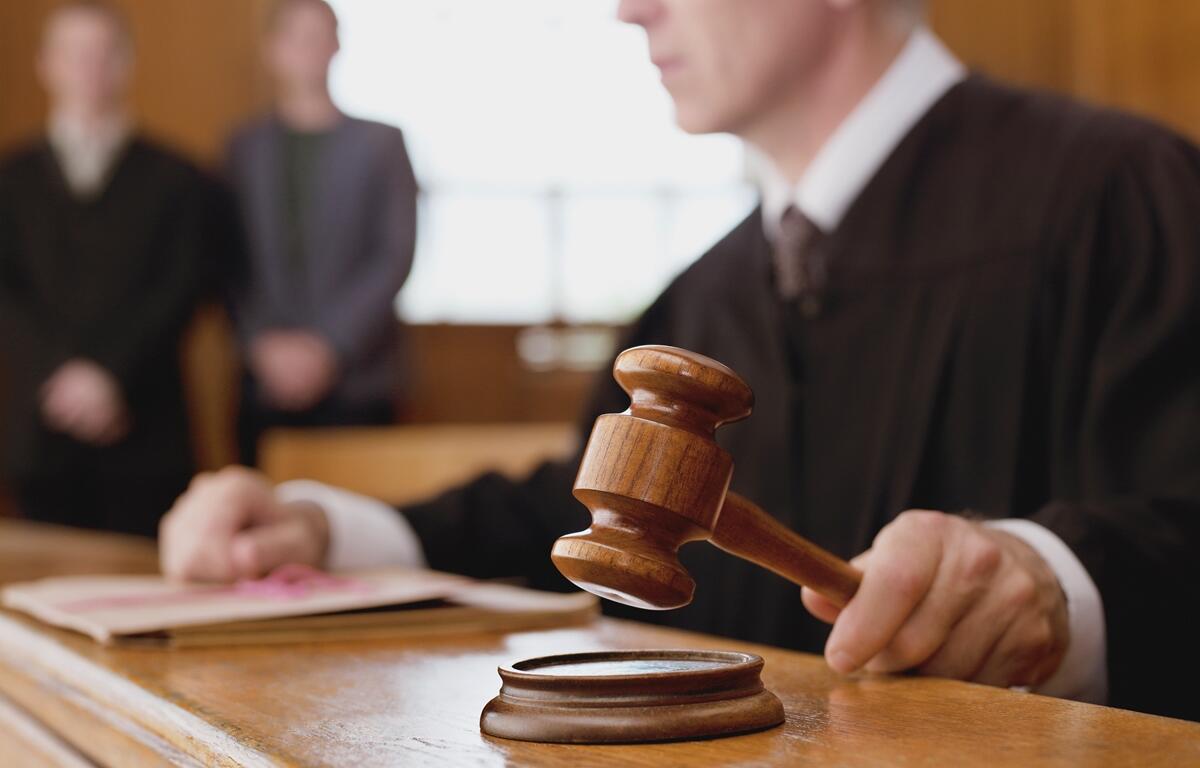Judge holding gavel in courtroom with two people standing in background.