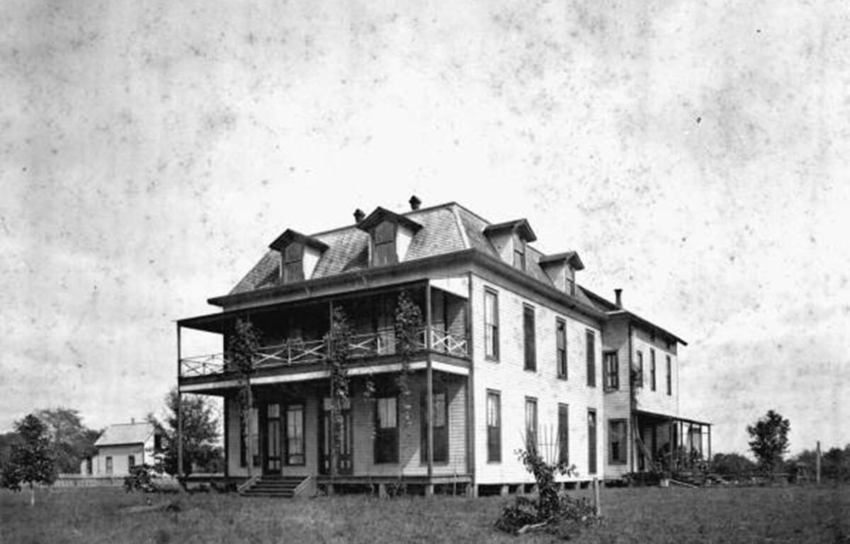 A historic black-and-white photograph of a large, two-story wooden house with a distinctive mansard roof featuring multiple dormer windows. The building has a wraparound porch on both levels, supported by slender columns with climbing vines. Tall windows line the sides of the structure. The house is elevated slightly above the ground and sits in an open grassy area with a few scattered trees and a smaller building visible in the background.