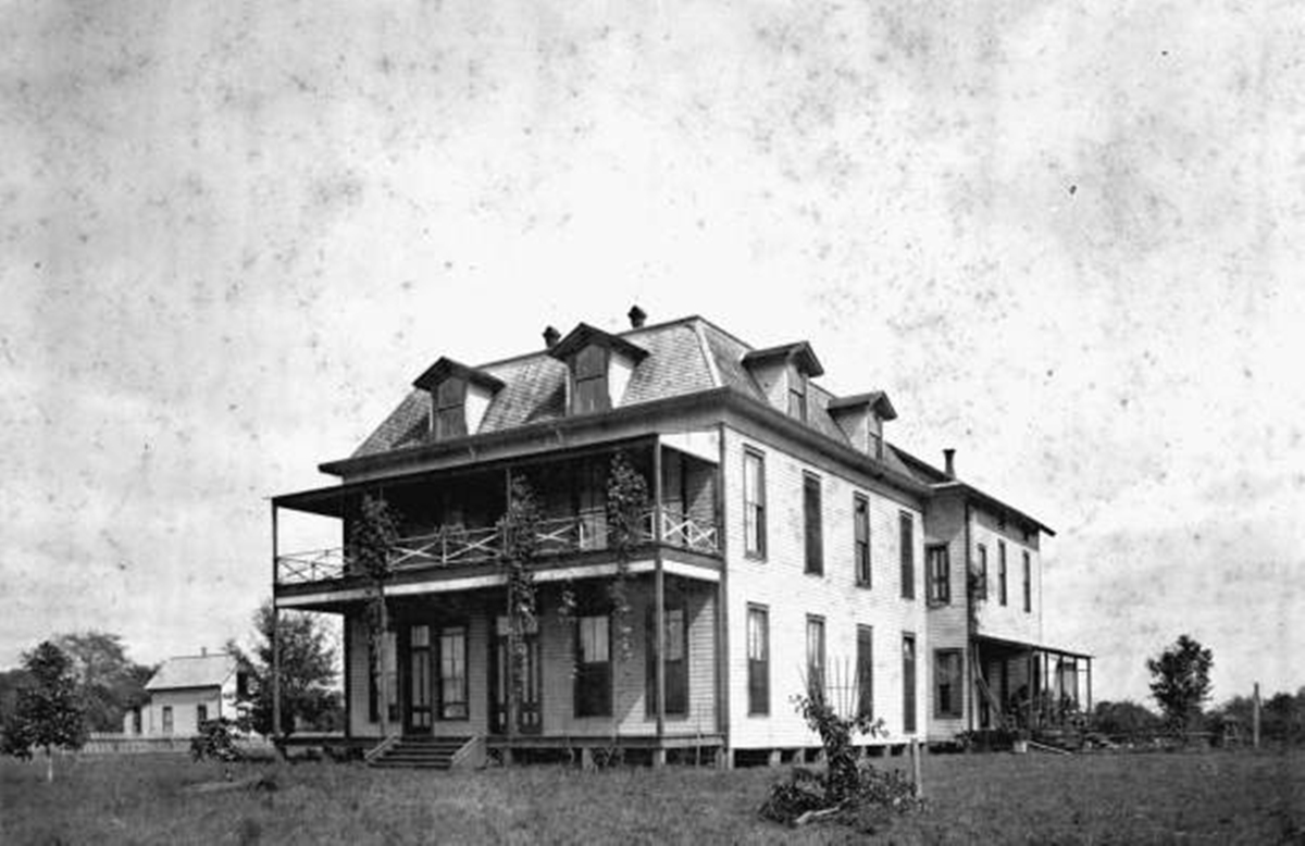 A historic black-and-white photograph of a large, two-story wooden house with a distinctive mansard roof featuring multiple dormer windows. The building has a wraparound porch on both levels, supported by slender columns with climbing vines. Tall windows line the sides of the structure. The house is elevated slightly above the ground and sits in an open grassy area with a few scattered trees and a smaller building visible in the background.