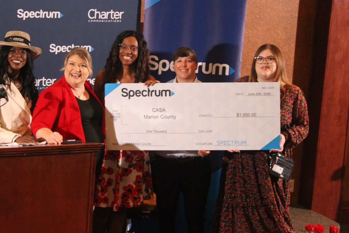 Five well-dressed women standing in front of a large white poster board, made to look like a giant check, with letter and numbers, in front of a blue background.