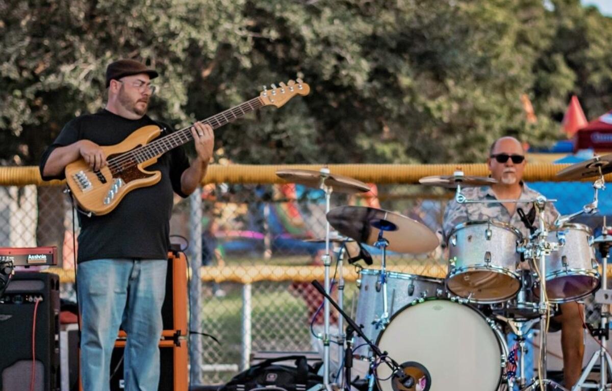Two musicians playing bass guitar and drums during an outdoor community event with playground equipment in the background.