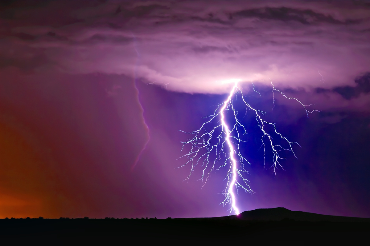 Lightning bolt flashes across the sky during a thunderstorm over a flat landscape at dusk.
