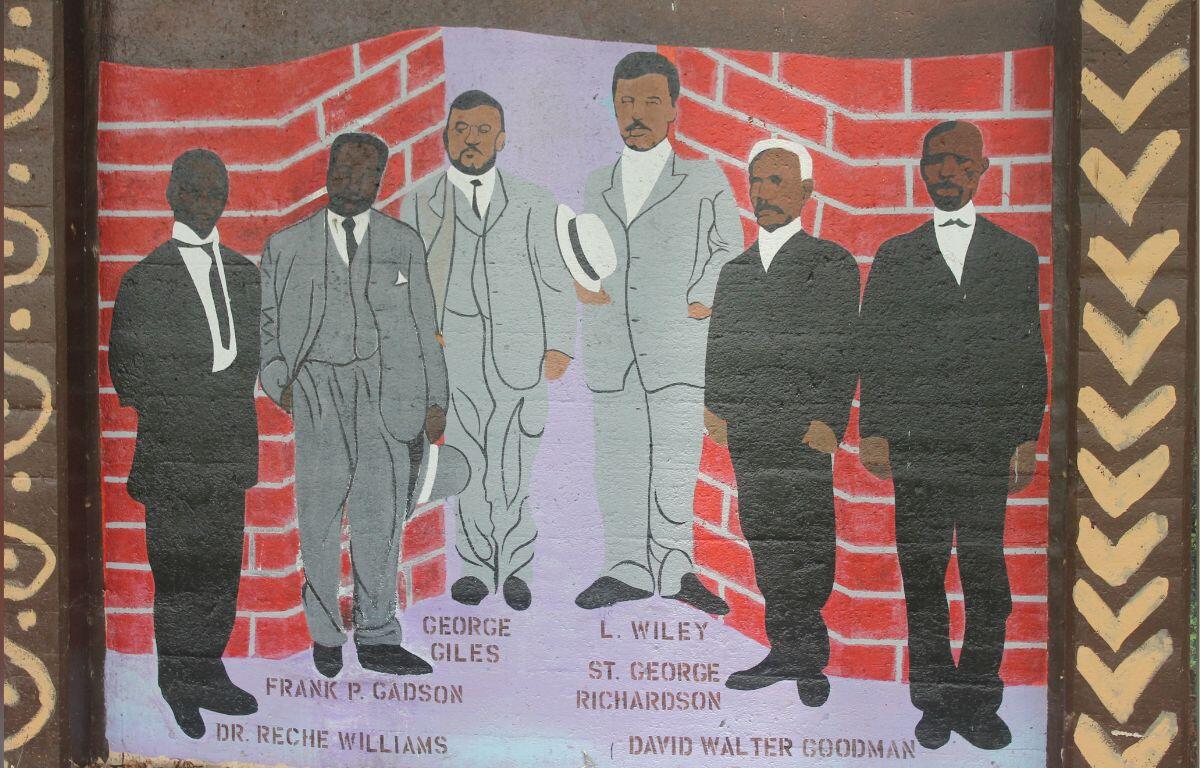 Six men in suits standing in front of brick wall, on top of a gray floor, with their names underneath them.