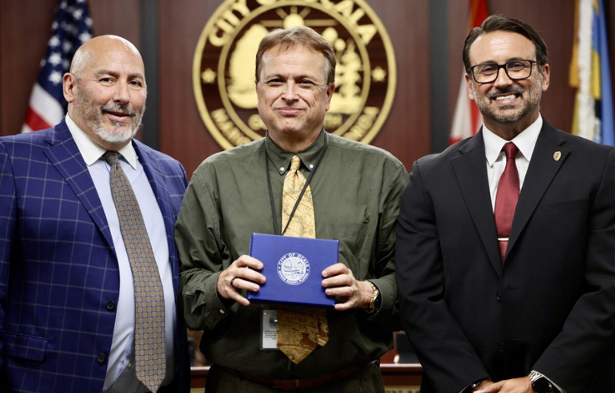 Three men standing in front of a flag and a circle with an insignia, with the man in the middle holding a box,