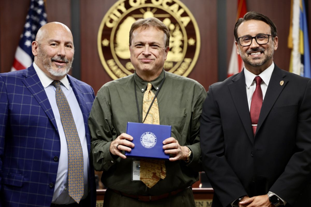 Three men standing in front of a flag and a circle with an insignia, with the man in the middle holding a box,