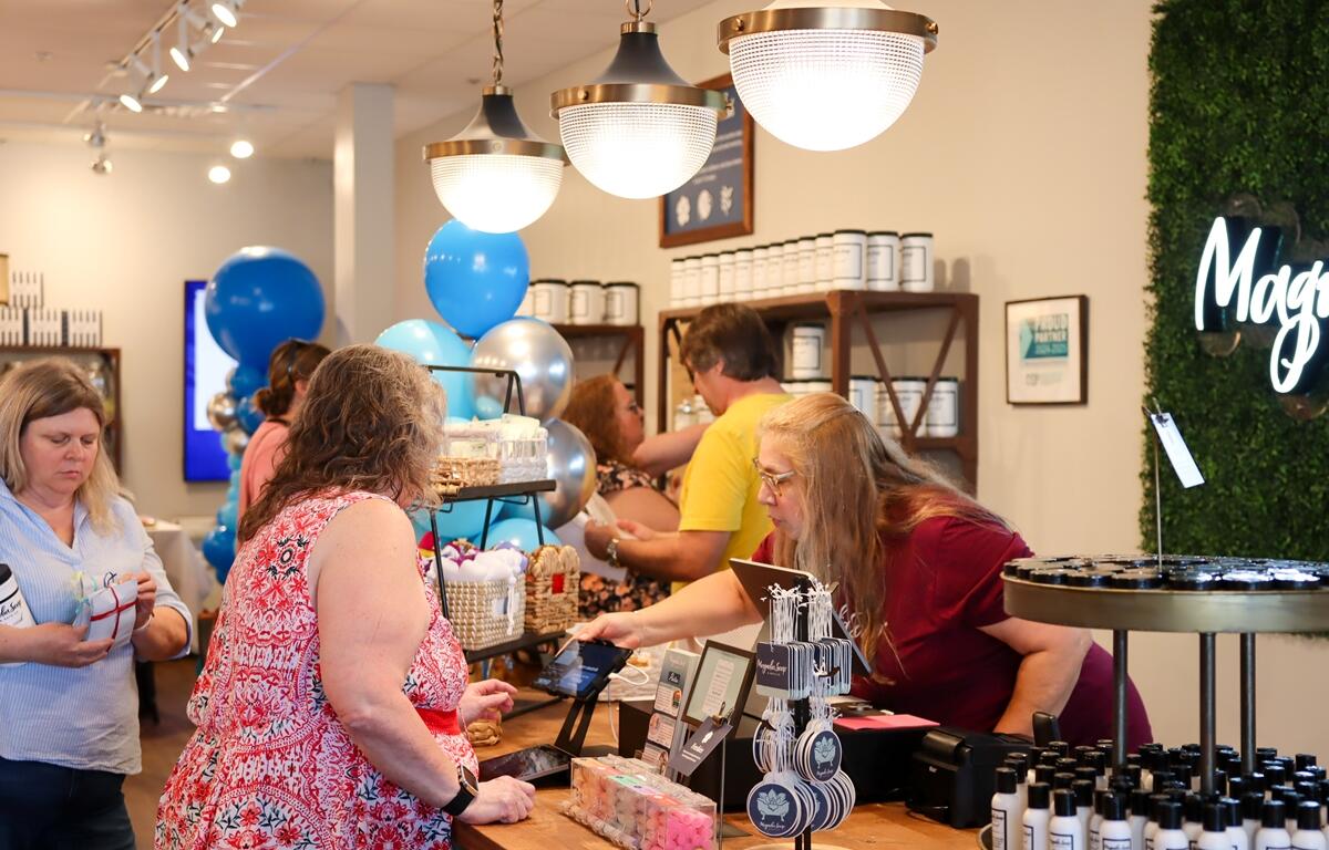 Shoppers at the checkout counter inside Magnolia Soap and Bath Company during its grand opening event, with blue balloons and product displays in the background.