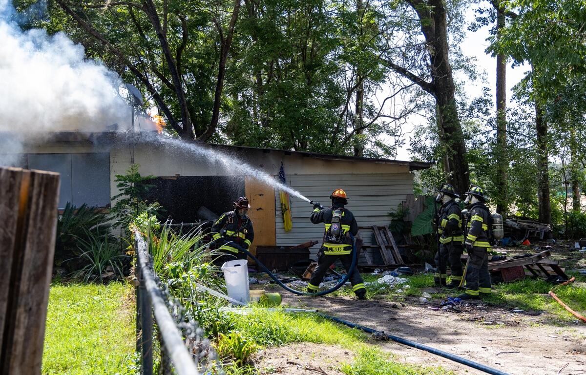 Firefighters use a hose to spray water on a burning house while smoke rises from the roof.