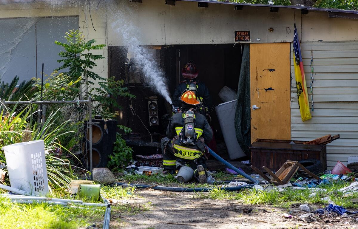 Two firefighters spray water into a smoke-damaged home in Ocala surrounded by debris and overgrown vegetation.